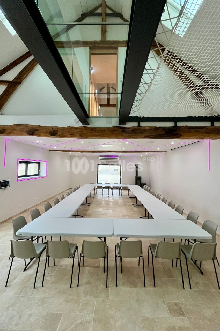 Salle de réunion lumineuse avec tables disposées en U, chaises alignées et poutres apparentes au plafond.