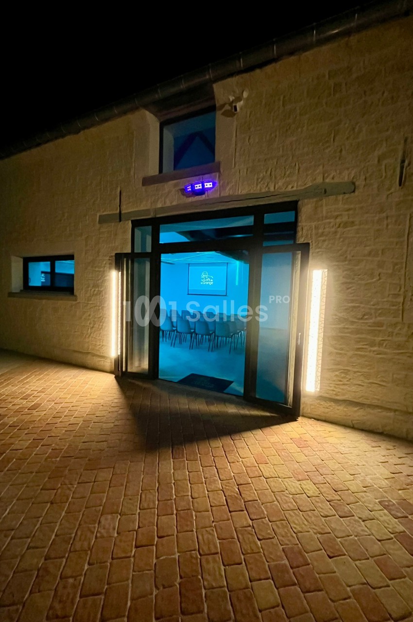 Entrée éclairée d'un bâtiment en pierre de nuit, avec une salle de conférence visible à travers les portes vitrées.