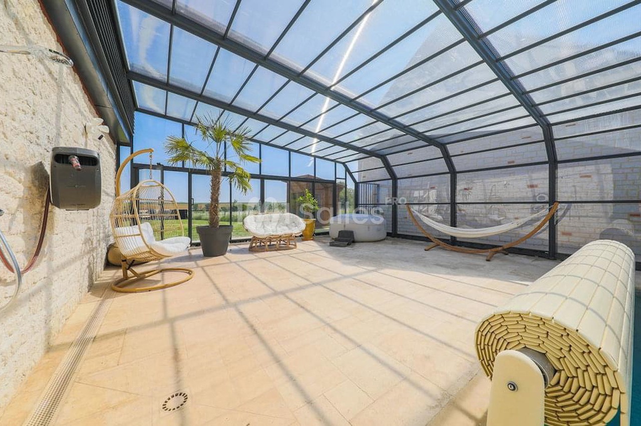 Véranda lumineuse avec piscine couverte, mobilier de détente, hamac et vue sur un jardin extérieur.