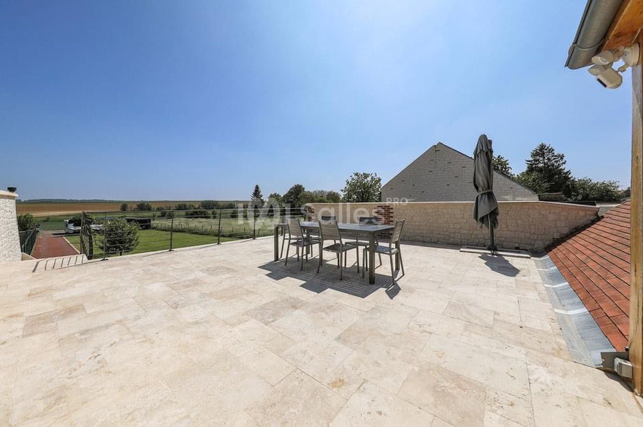 Terrasse spacieuse en pierre avec table, chaises et parasol, offrant une vue dégagée sur la campagne environnante.