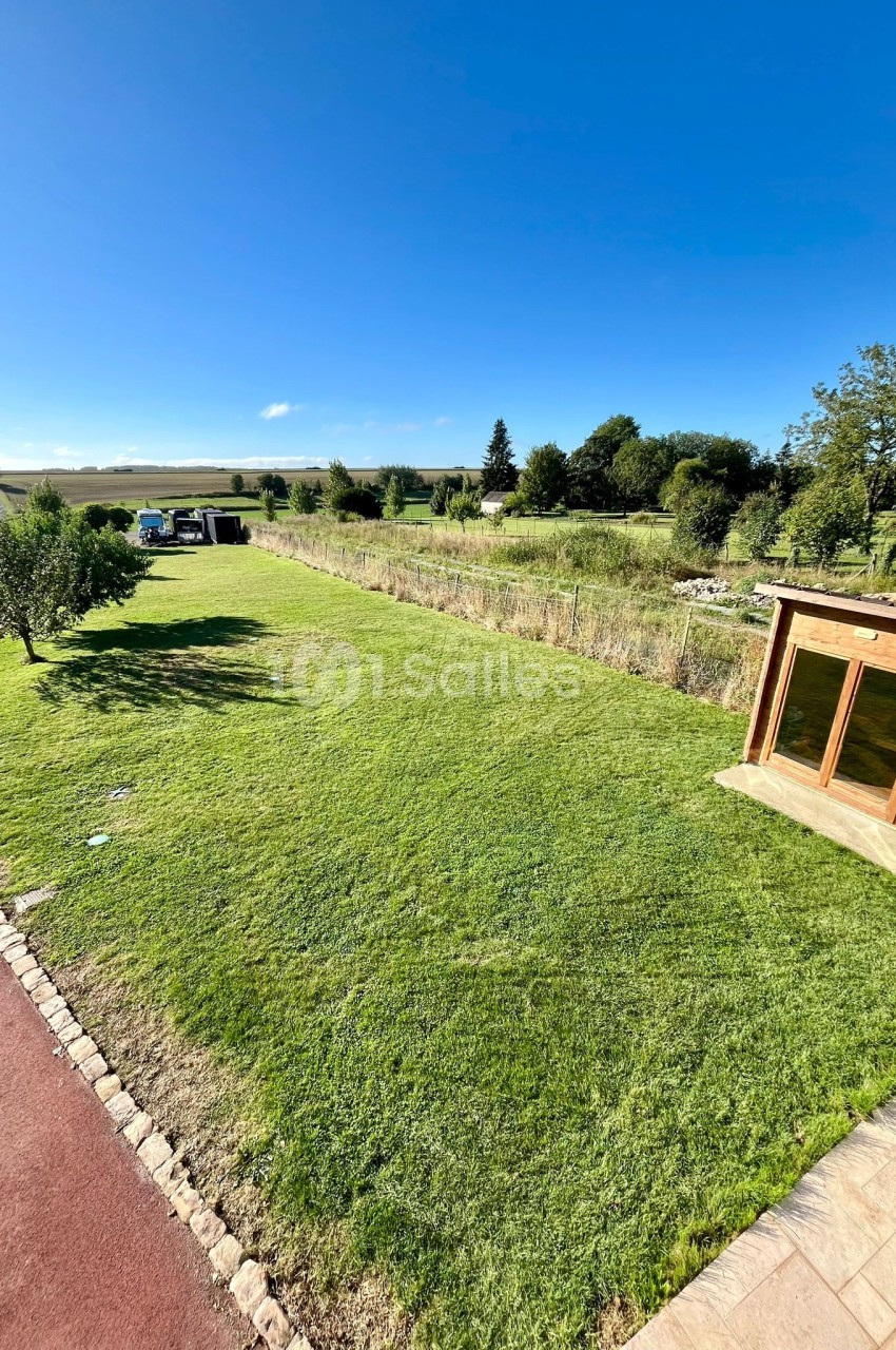 Jardin avec pelouse bien entretenue, chemin pavé, cabanon en bois et paysage rural en arrière-plan sous un ciel bleu.