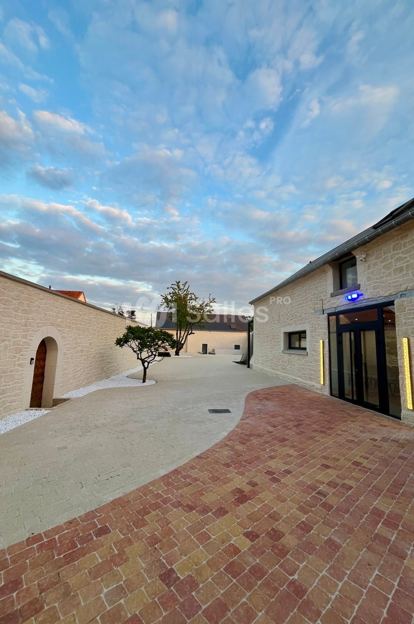 Cour pavée avec un arbre central, entourée de bâtiments en pierre claire sous un ciel partiellement nuageux.