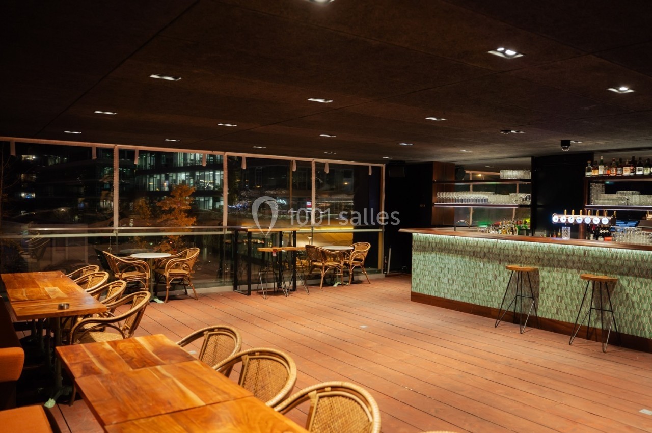 Salle de restaurant moderne avec tables en bois, chaises en rotin et bar éclairé, vue sur la ville à travers de grandes…