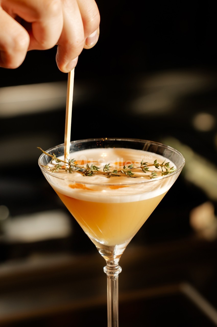 Un cocktail jaune dans un verre à martini décoré de thym et d'une mousse blanche, une main ajustant une garniture.