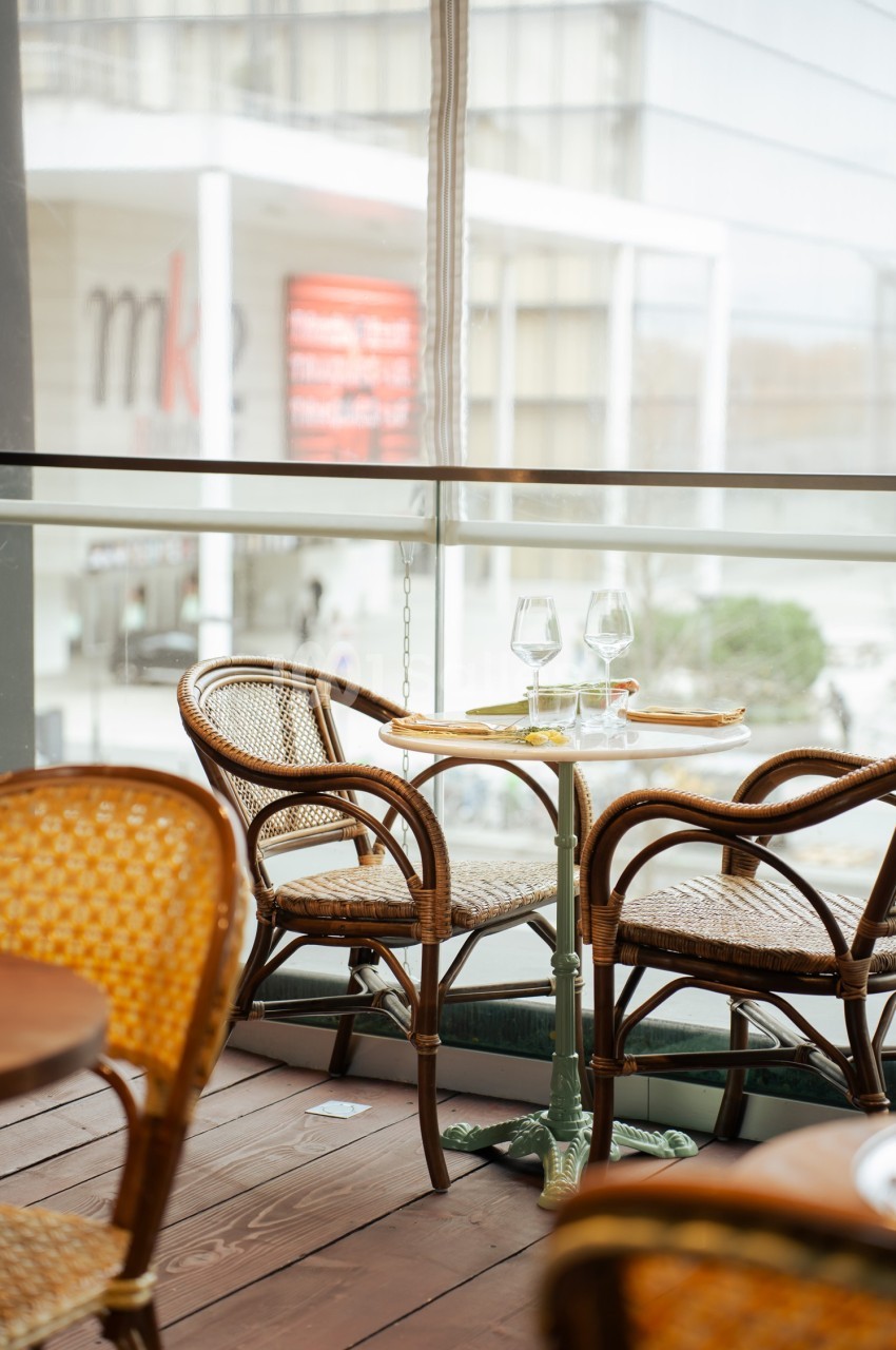 Table en terrasse avec chaises en rotin, deux verres à vin et assiettes, vue sur un bâtiment moderne à travers une baie…