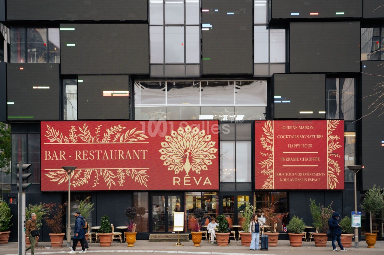Façade d'un bar-restaurant avec terrasse, enseignes rouges affichant le nom ’Réva’ et des informations sur les services.