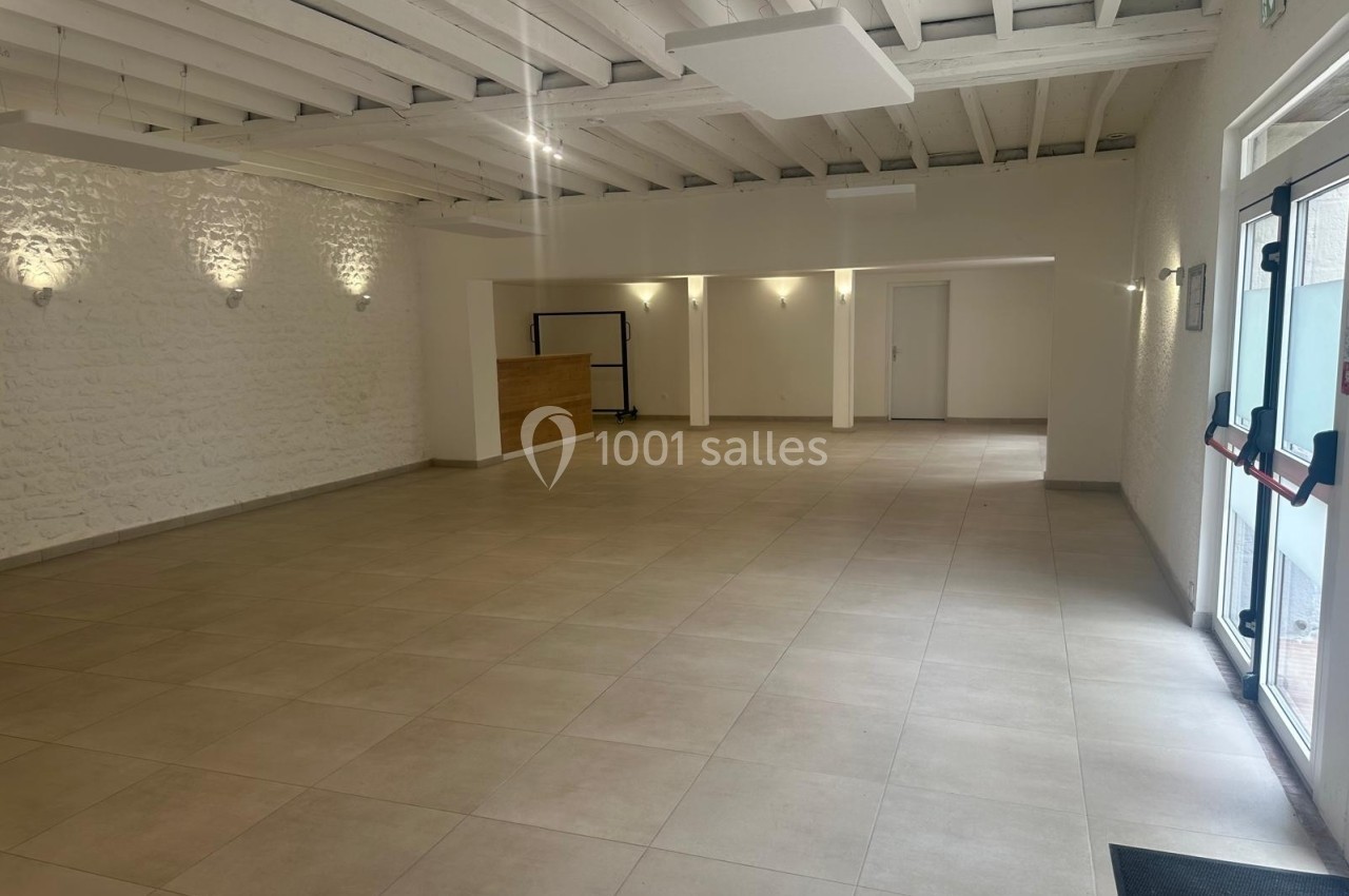 Salle vide lumineuse avec murs blancs, sol carrelé beige, plafond avec poutres apparentes et porte vitrée à droite.