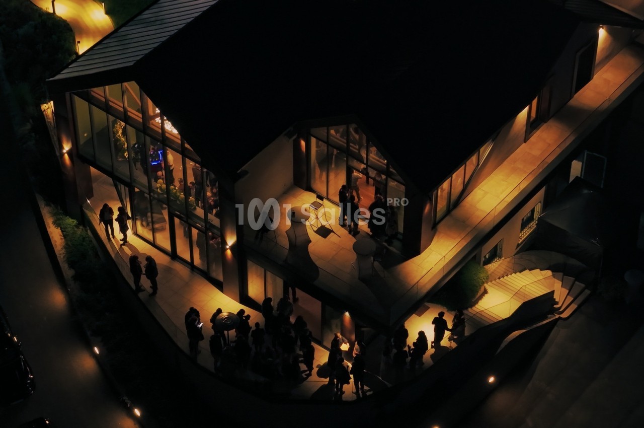 Vue aérienne nocturne d'un bâtiment moderne éclairé avec des personnes rassemblées à l'extérieur et à l'intérieur.
