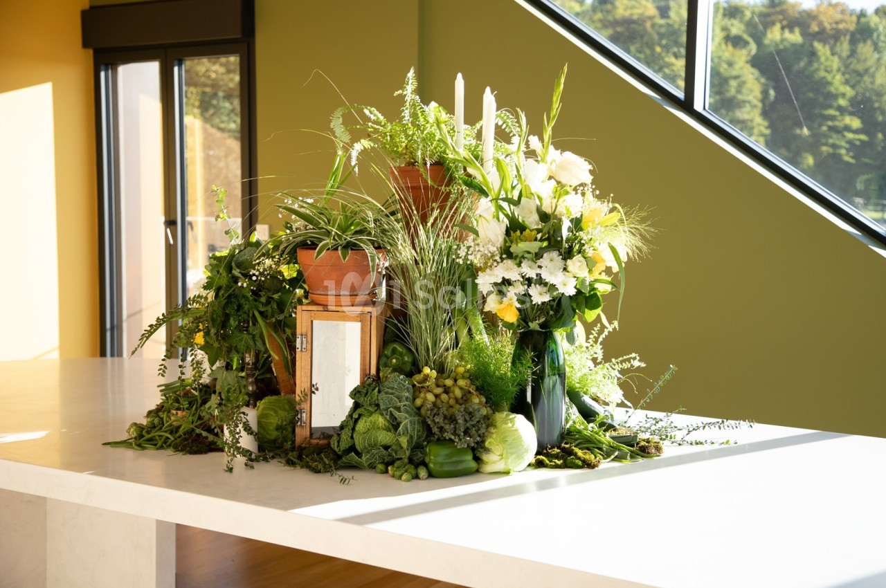 Arrangement de plantes vertes, fleurs blanches et légumes disposés sur une table blanche près d'une fenêtre.