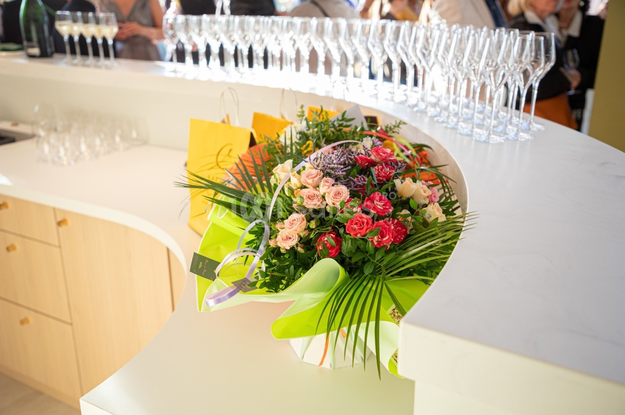 Bouquet de fleurs colorées posé sur un comptoir blanc, entouré de verres à pied et de sacs cadeaux jaunes.