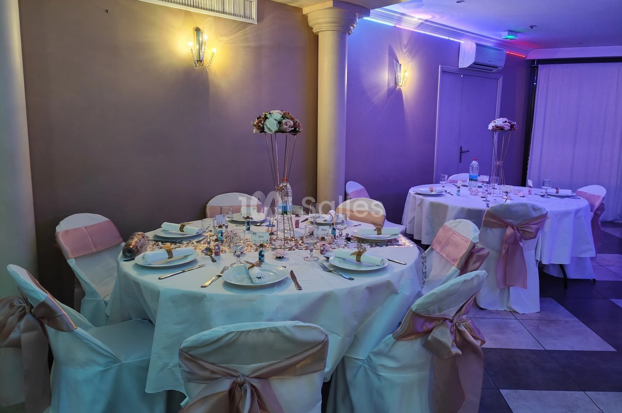 Salle de réception avec tables rondes décorées de nappes blanches, chaises ornées de nœuds roses et centres de table fleuris.