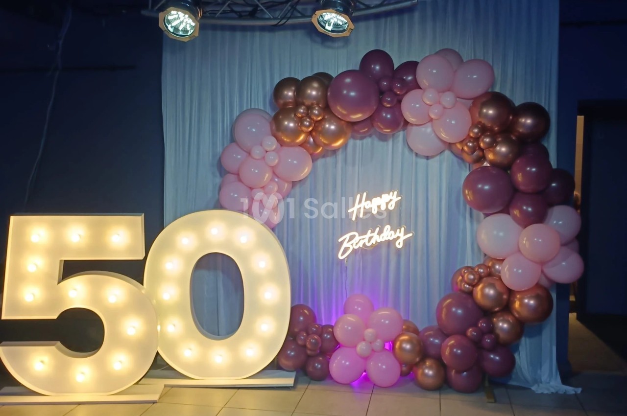 Décoration d'anniversaire avec un ’50’ lumineux, un arc de ballons roses et dorés, et un néon ’Happy Birthday’.