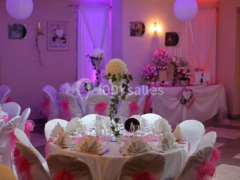 Salle décorée pour un mariage avec des tables dressées, des chaises ornées de nœuds roses et des éclairages colorés.