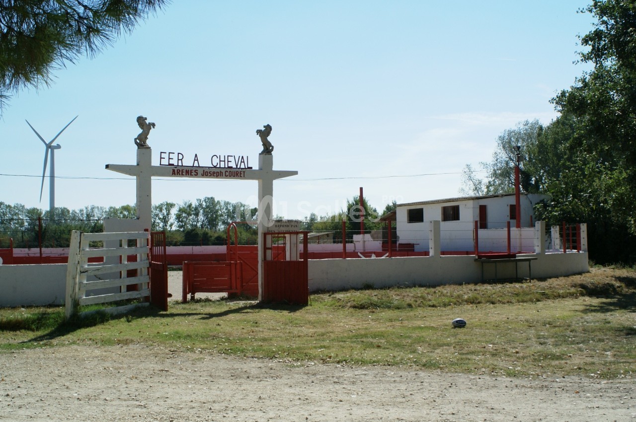 Entrée des arènes Joseph Coudert avec portail blanc et rouge, entourée de végétation et d'une éolienne en arrière-plan.