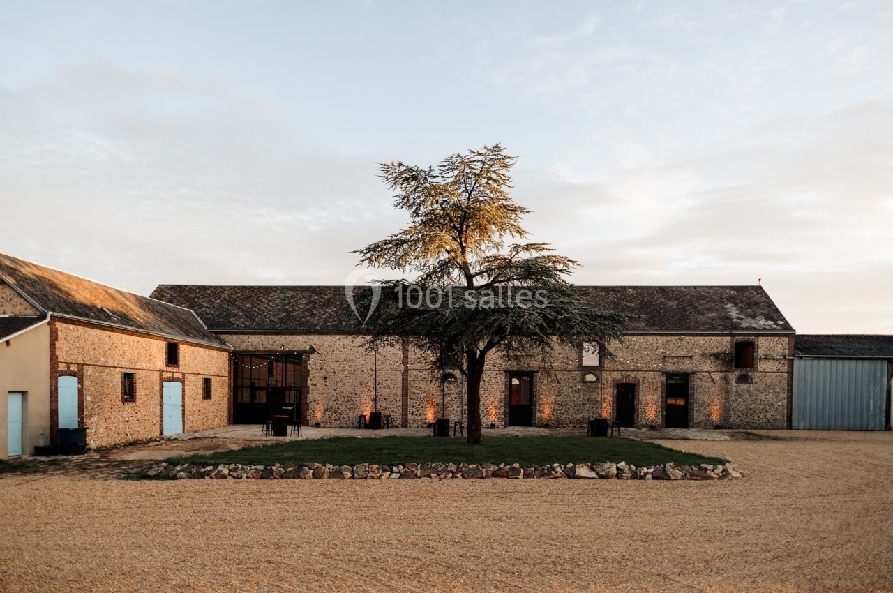 Bâtiment en pierre avec toit en ardoise, entouré de gravier et d'un arbre central sous un ciel dégagé.