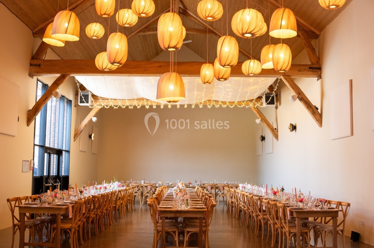 Grande salle de réception décorée avec des tables alignées, des chaises en bois et des suspensions lumineuses en rotin.