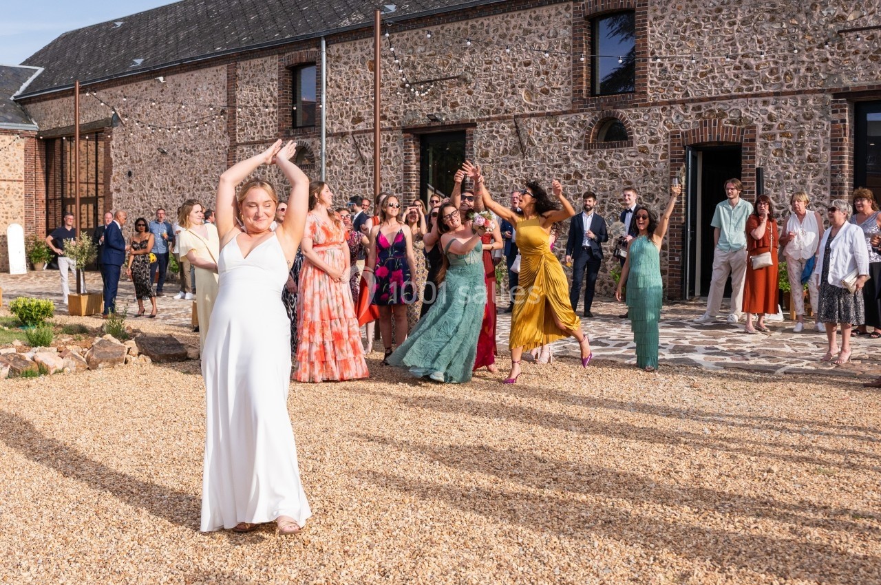 Une femme en robe blanche lance un bouquet devant un groupe de personnes lors d'une réception en extérieur.