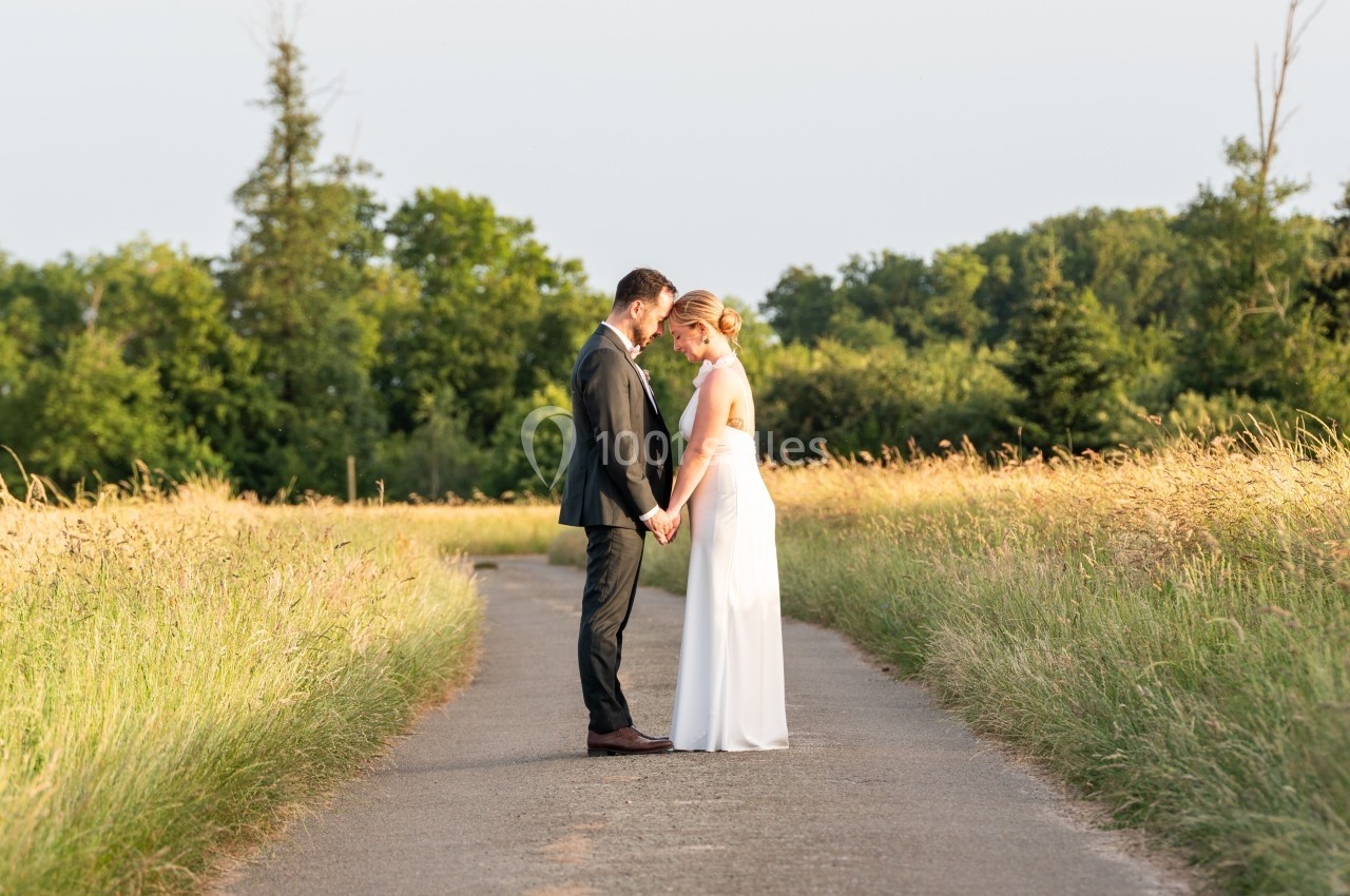 Un couple en tenue de mariage se tient par les mains au milieu d'un chemin bordé d'herbes hautes, sous un ciel dégagé.