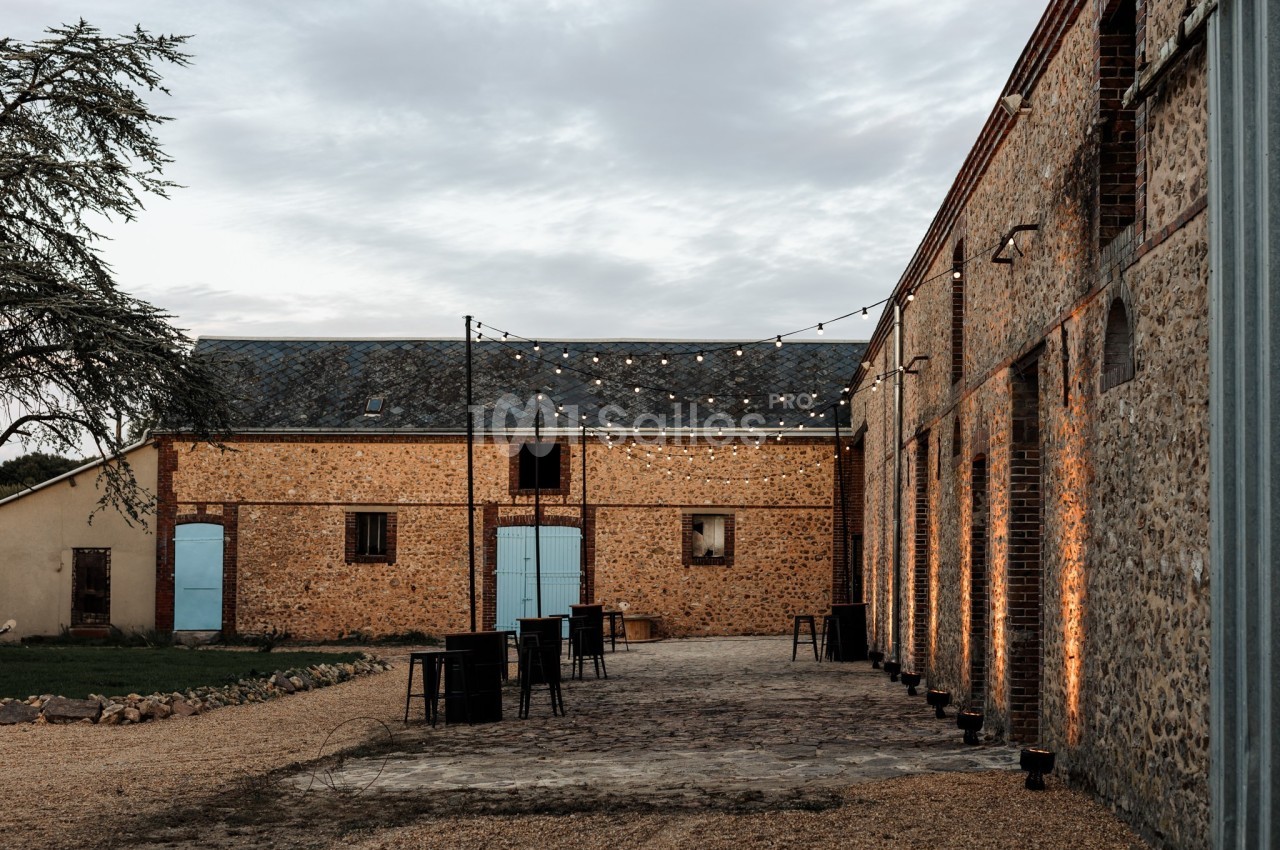 Cour extérieure d'une bâtisse en pierre avec guirlandes lumineuses suspendues et tables hautes disposées.