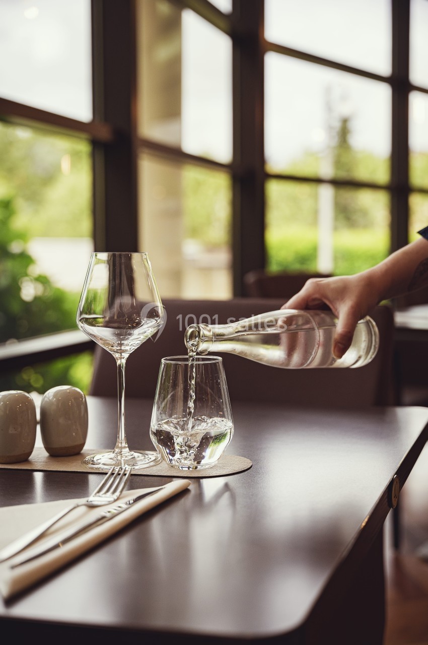 Une main verse de l'eau dans un verre sur une table dressée avec des couverts et des verres dans un restaurant lumineux.