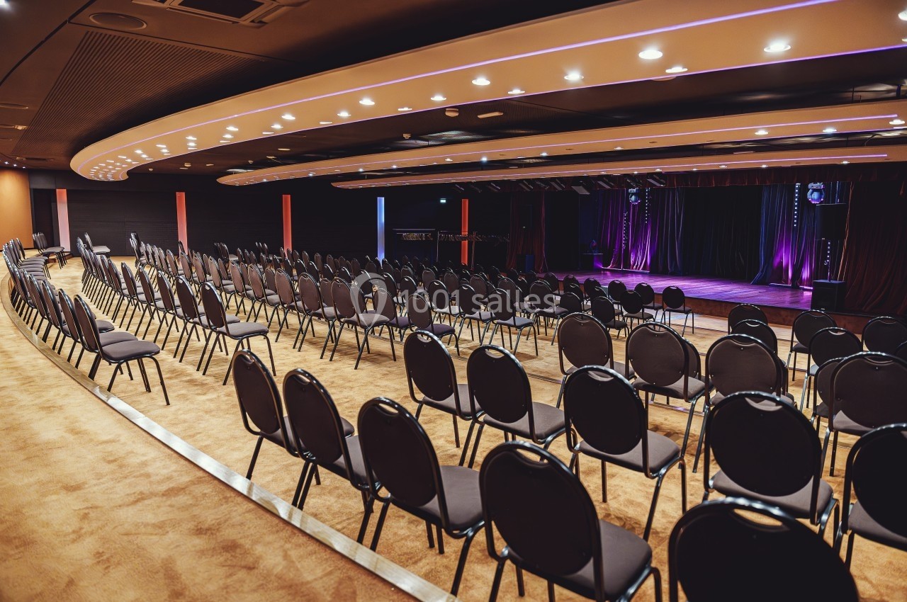 Salle de conférence vide avec des rangées de chaises noires alignées face à une scène éclairée par des projecteurs.