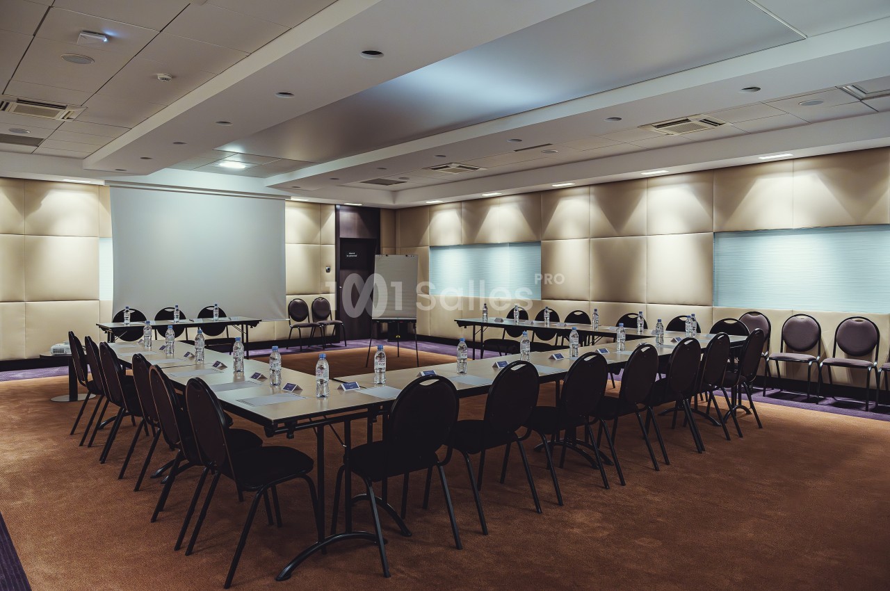 Salle de réunion équipée de tables disposées en U, chaises alignées et éclairage tamisé.