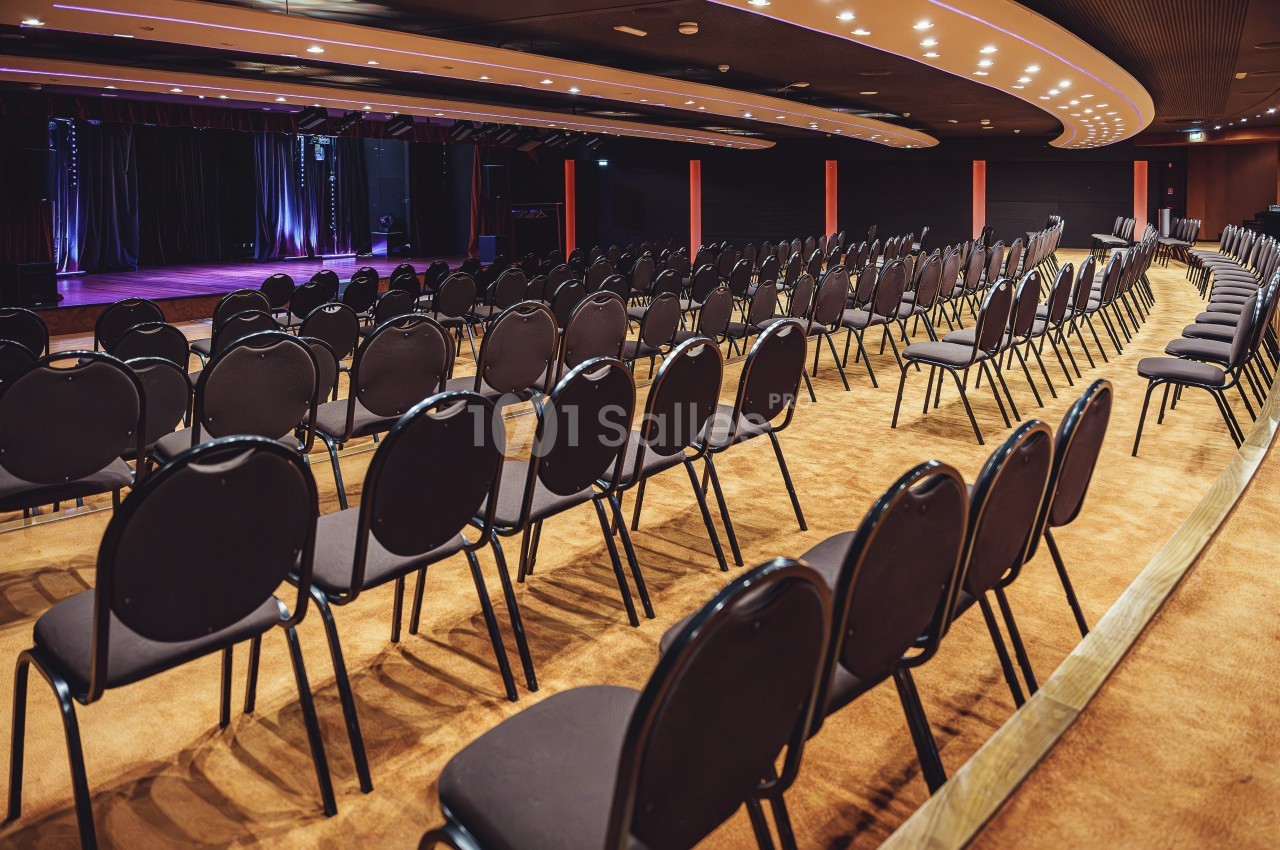 Salle de spectacle vide avec des rangées de chaises noires alignées face à une scène éclairée.