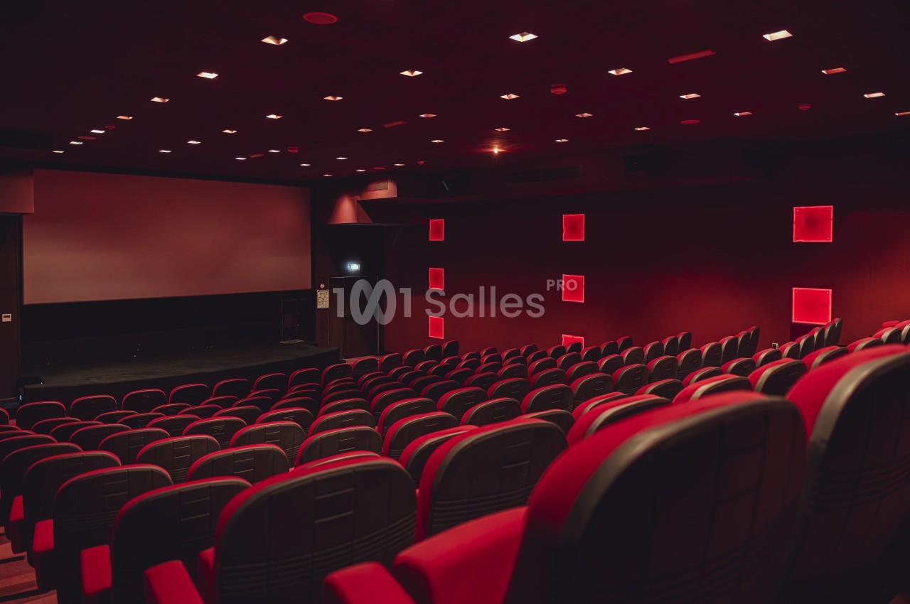 Salle de cinéma vide avec rangées de fauteuils rouges, écran blanc et éclairage tamisé.