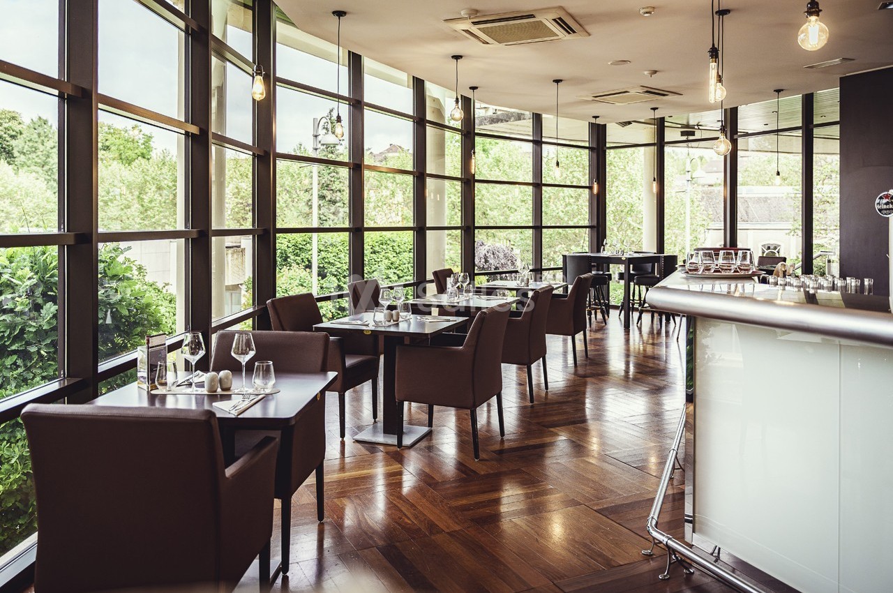 Salle de restaurant lumineuse avec grandes baies vitrées, tables dressées et mobilier moderne en bois et cuir.