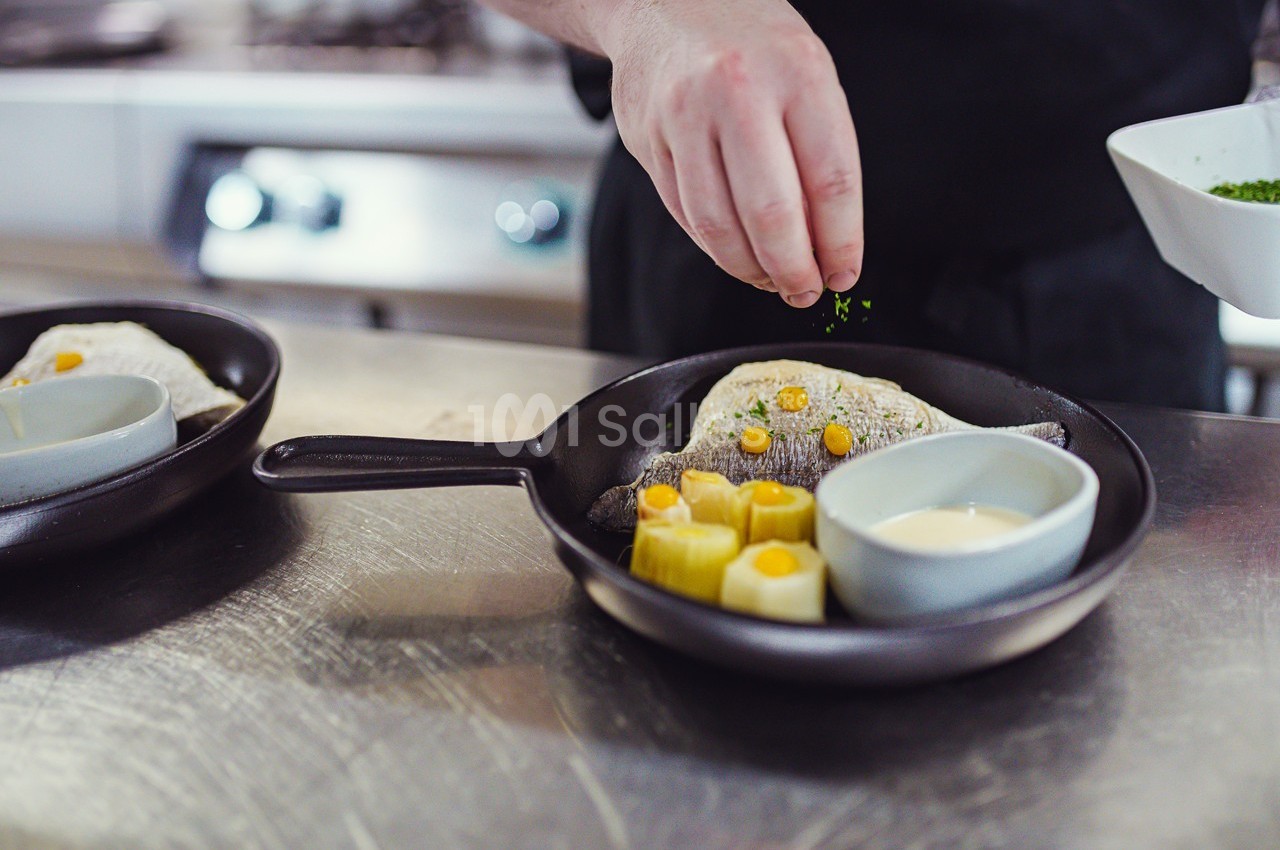 Un chef saupoudre des herbes sur un plat contenant du poisson, des légumes et une sauce, présenté dans une poêle.