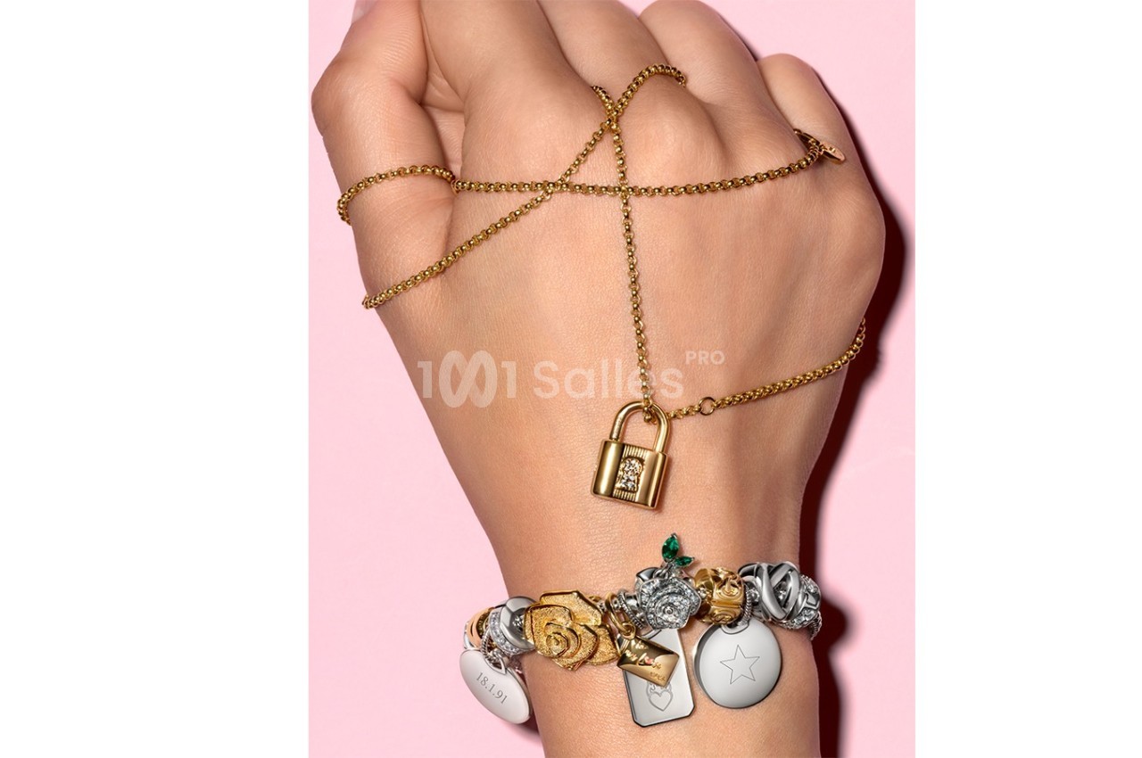 Main portant un bracelet orné de charms variés et tenant une chaîne dorée avec un pendentif en forme de cadenas.