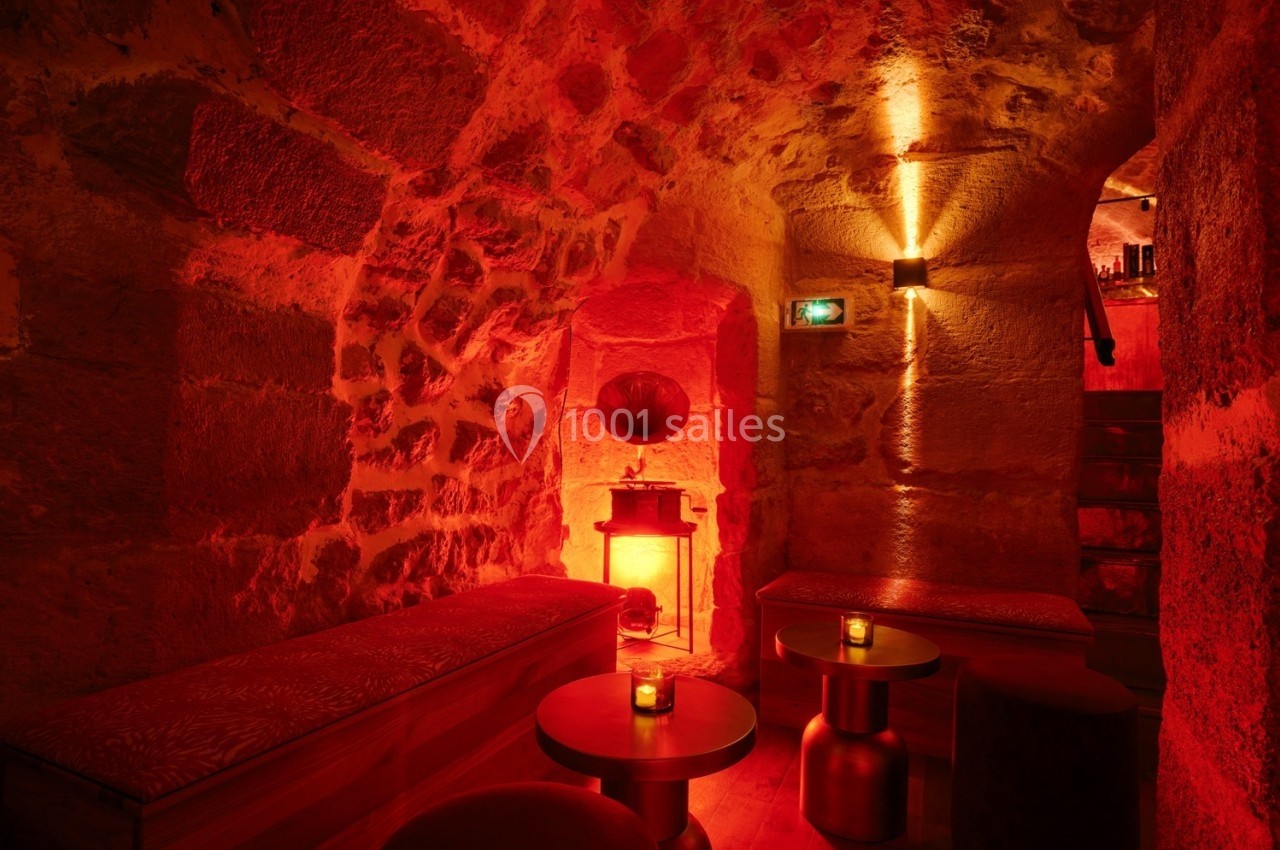 Salle voûtée en pierre éclairée par une lumière rouge tamisée, avec banquettes, tables basses et bougies.
