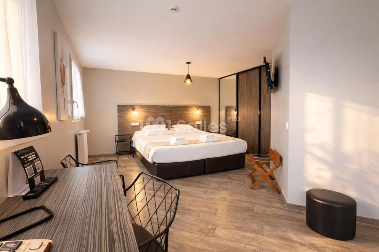 Chambre d'hôtel moderne avec deux lits simples, bureau, chaise, télévision murale et éclairage chaleureux.