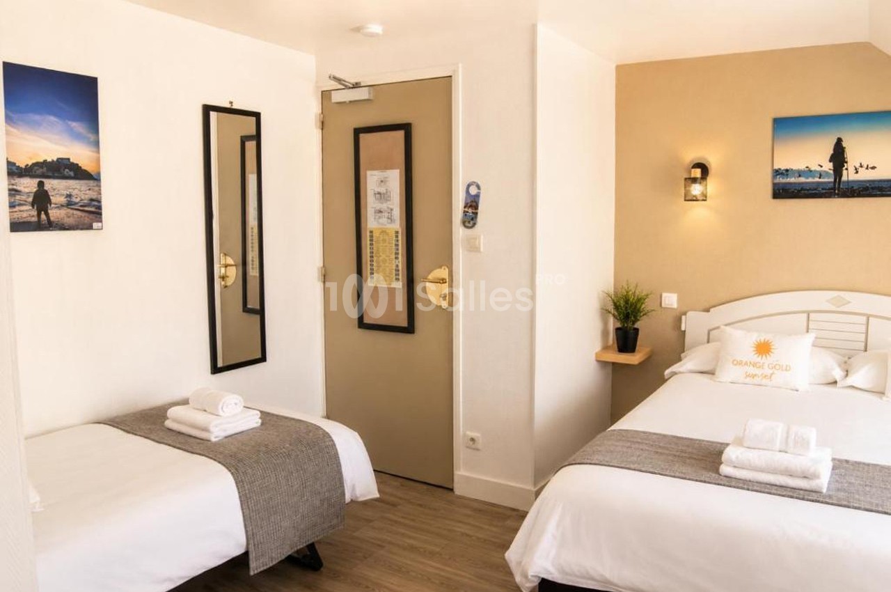 Chambre d'hôtel avec deux lits, décor sobre, murs clairs, tableaux, miroir, et porte beige avec des informations affichées.