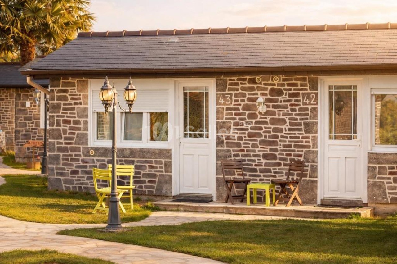 Façade d'un bâtiment en pierre avec portes blanches, mobilier de jardin coloré et allée en pierre sous une lumière douce.