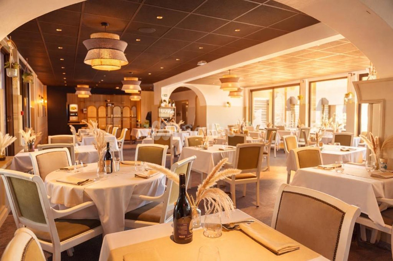 Salle de restaurant élégante avec tables dressées, chaises rembourrées et lumière naturelle entrant par de grandes fenêtres.