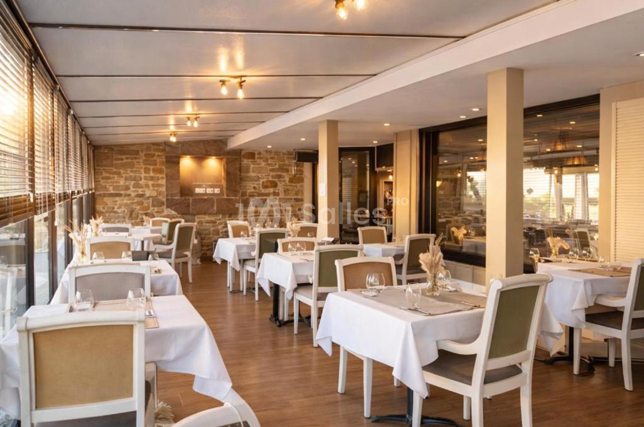 Salle de restaurant lumineuse avec tables dressées, chaises rembourrées et murs en pierre apparente.
