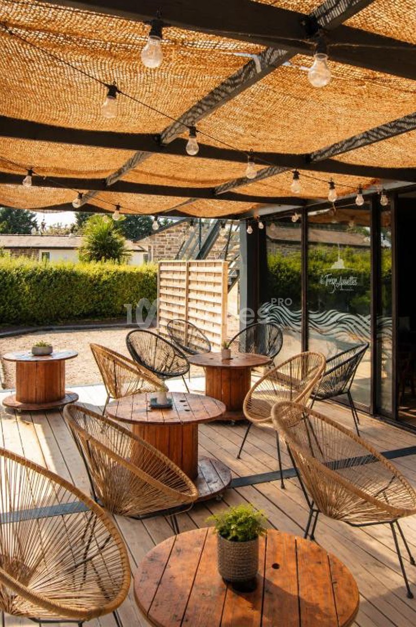 Terrasse en bois avec mobilier en rotin, tables rondes et toit en cannage laissant passer la lumière naturelle.