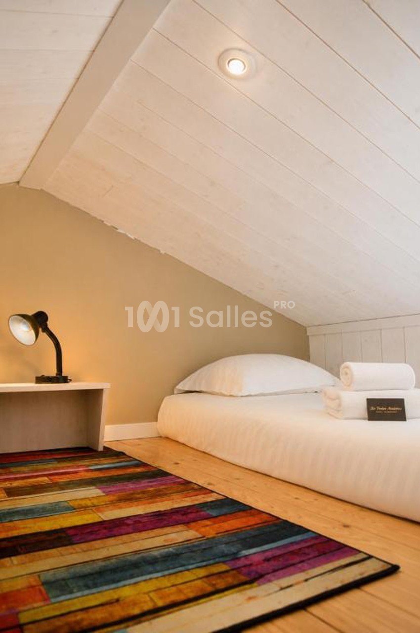 Lit simple avec draps blancs sous un plafond mansardé, lampe de chevet allumée et tapis coloré au sol.