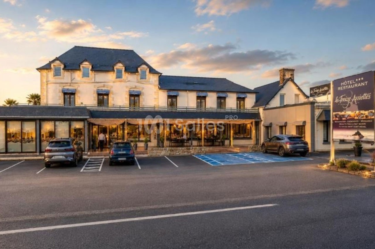 Façade d'un hôtel-restaurant avec parking, terrasse et enseigne visible sous un ciel partiellement nuageux.