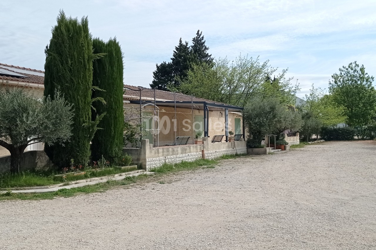 Allée gravillonnée bordée d'arbres et de buissons, longeant une maison avec terrasse et panneaux solaires.