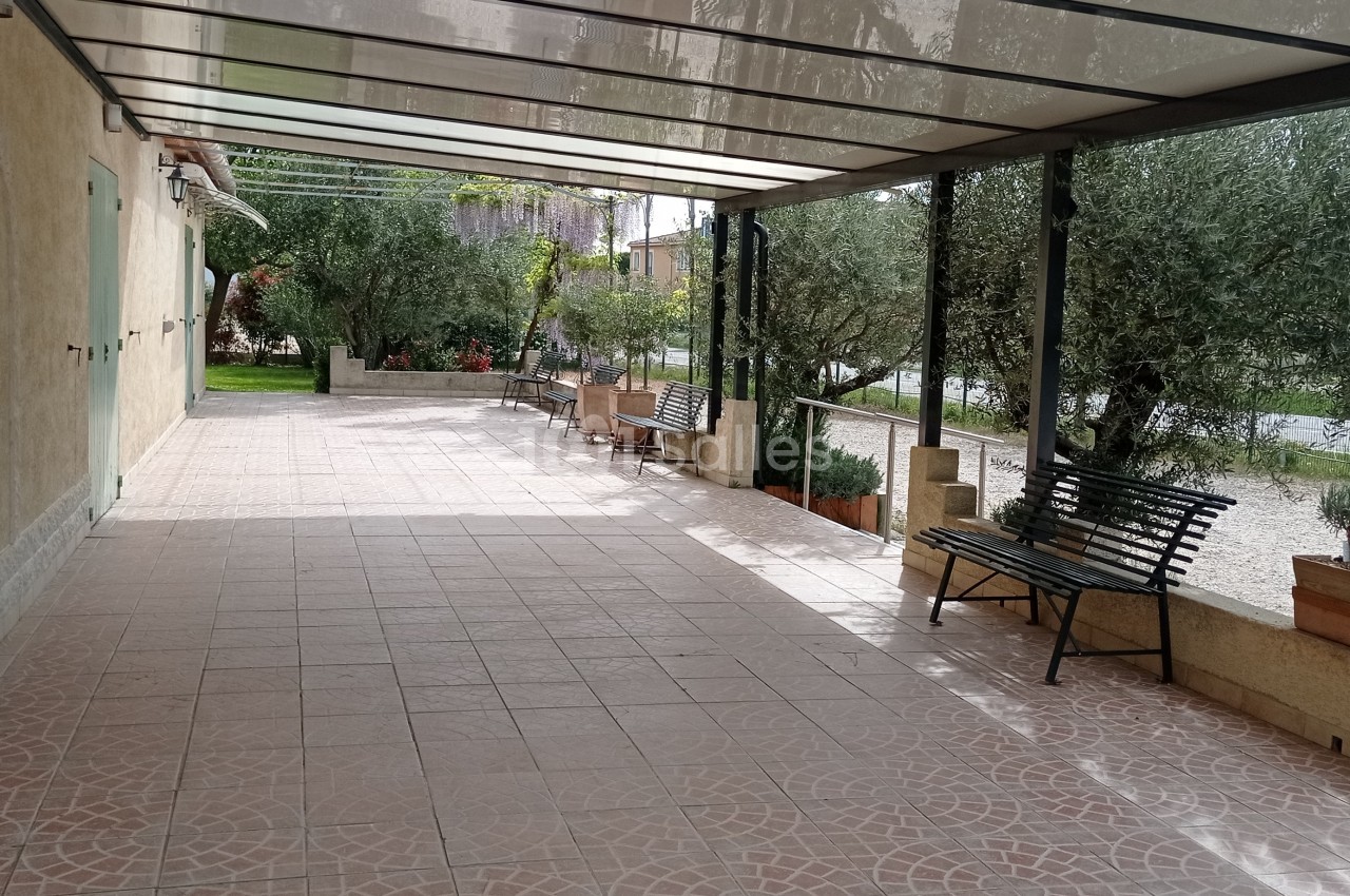 Terrasse couverte avec bancs en métal, sol carrelé et vue sur un jardin avec des oliviers.