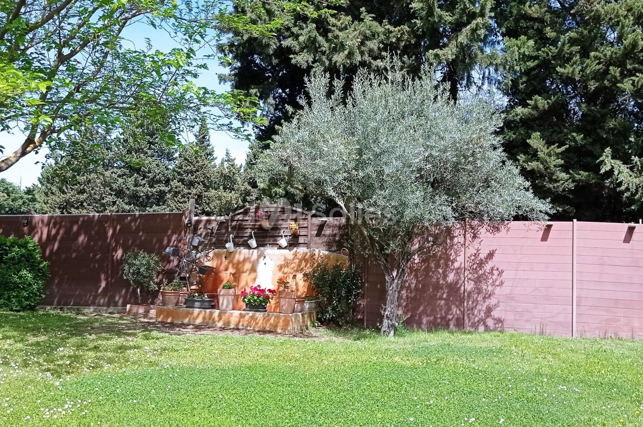 Un jardin avec pelouse, arbre, olivier, clôture en bois et un petit autel décoré de fleurs rouges.