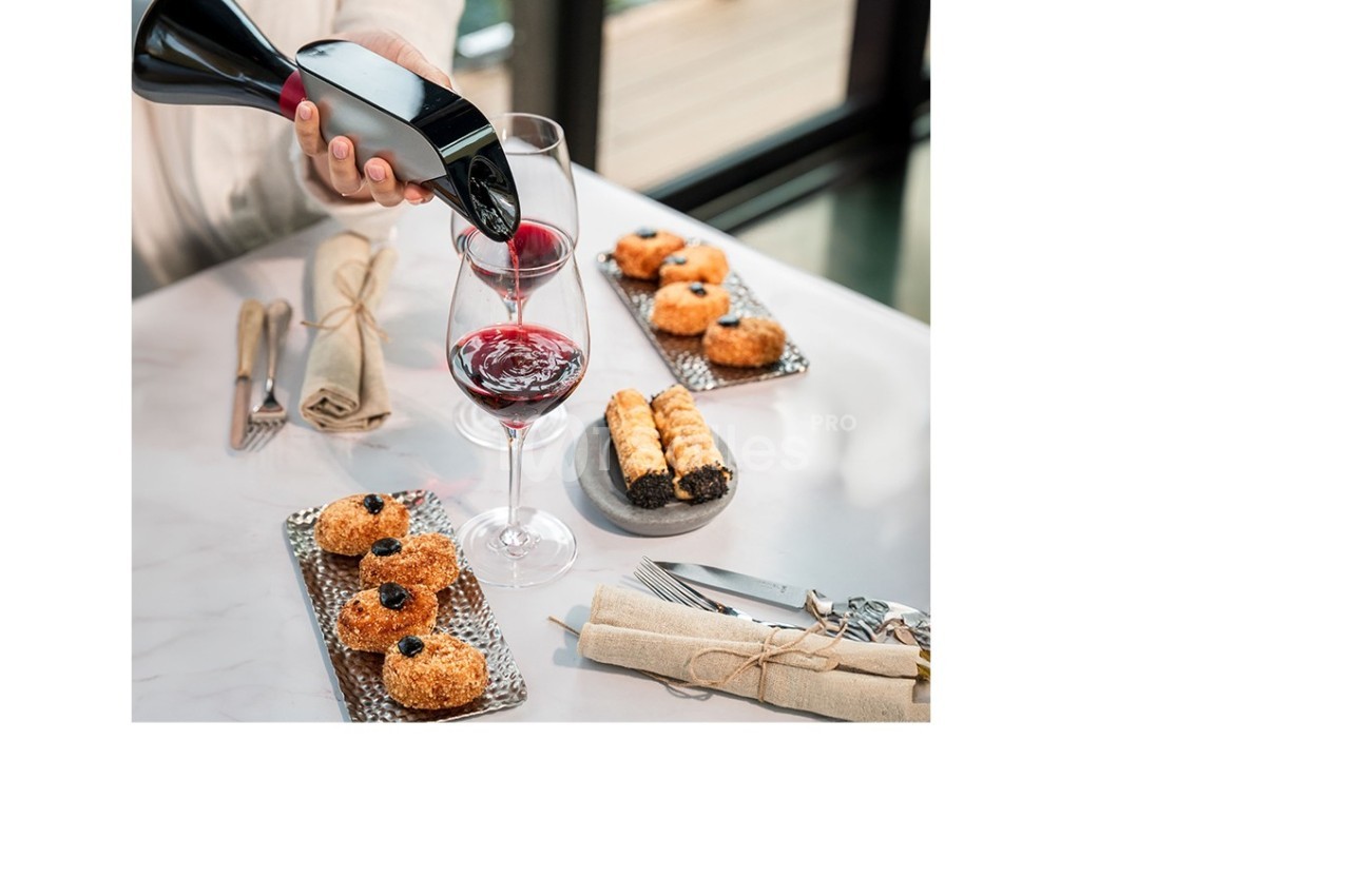 Une personne verse du vin rouge dans un verre sur une table dressée avec des pâtisseries et des couverts.