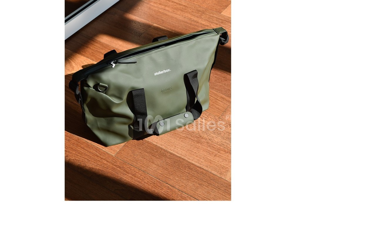 Sac vert en toile imperméable avec poignées noires, posé sur un sol en bois éclairé par la lumière naturelle.