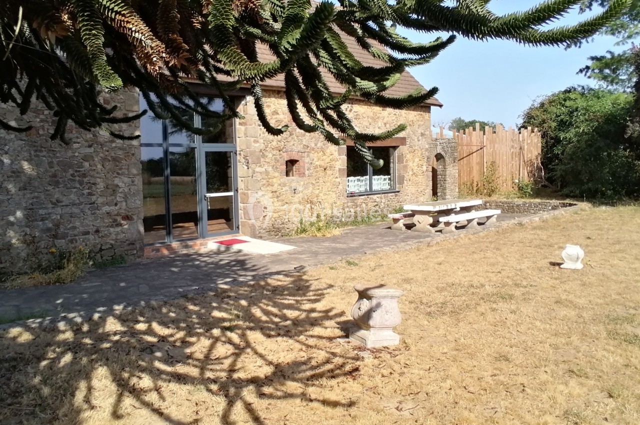 Façade en pierre d'une maison avec une table de jardin en pierre, entourée de pelouse sèche et d'arbres.