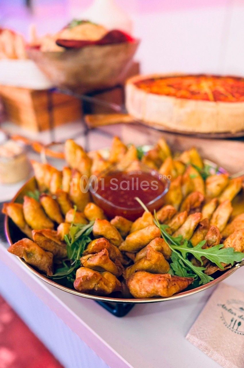 Assortiment de samoussas dorés avec sauce rouge au centre, décoré de roquette, présenté sur une table de buffet.