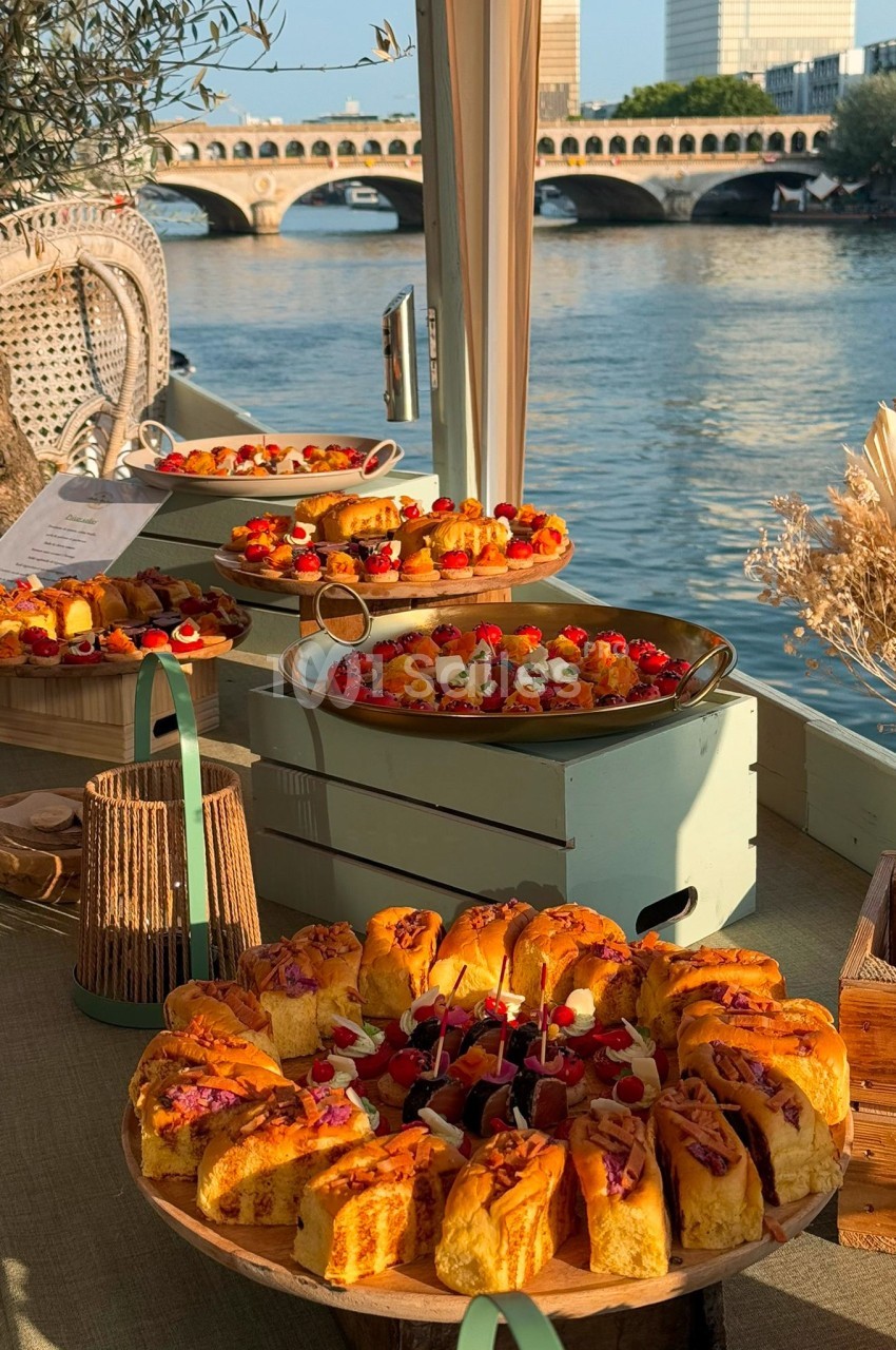 Plateaux de pâtisseries variées disposés sur une table en bois, avec vue sur une rivière et un pont en arrière-plan.