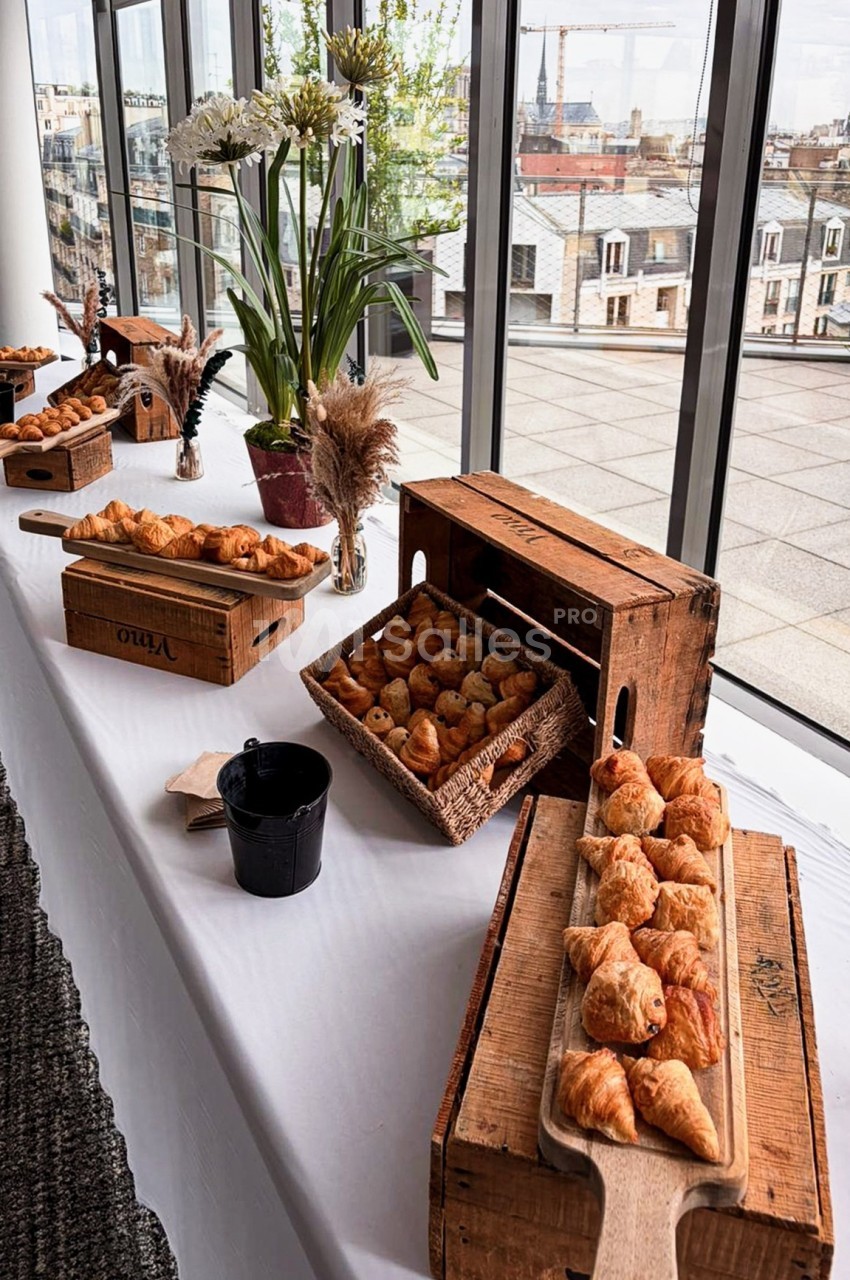 Table dressée avec des viennoiseries sur des planches en bois, entourée de décorations florales, avec vue sur des toits…