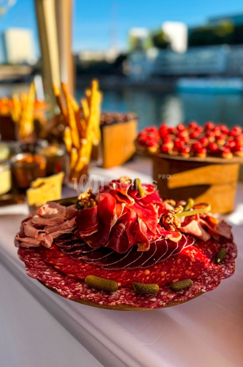 Plateau de charcuterie avec tranches de jambon, saucisson, cornichons et noix, disposé sur une table en extérieur.
